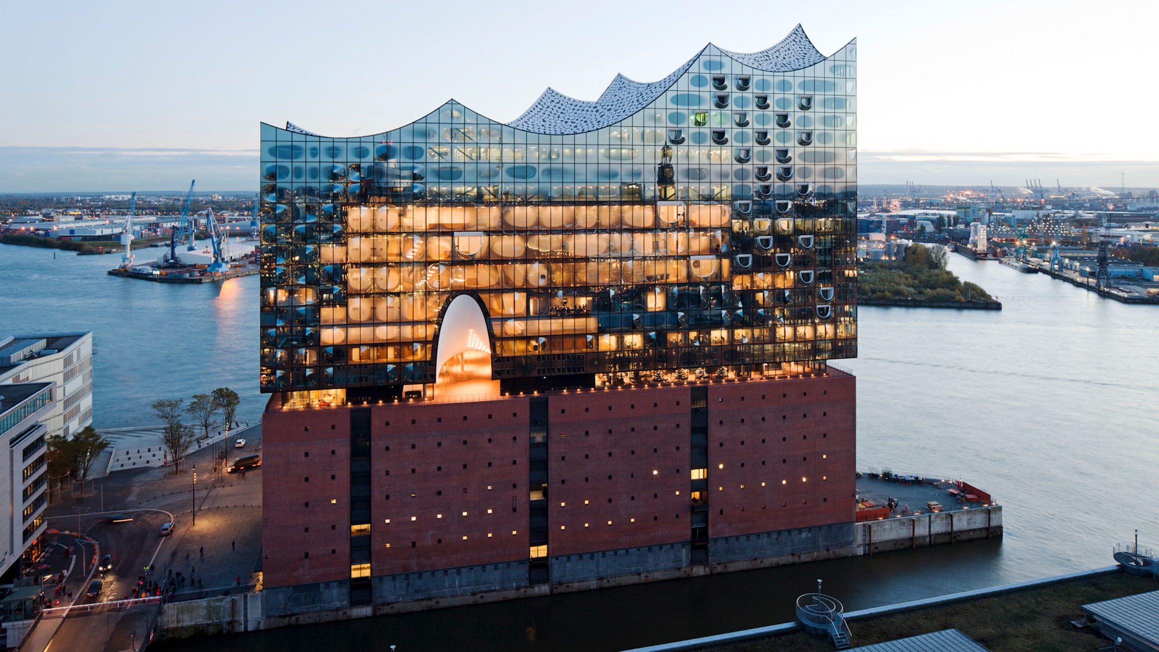 Elbphilharmonie in Hamburg, modernes Glasgebäude auf historischem Backsteinsockel bei Sonnenuntergang, mit Blick auf den Hafen und die Stadt im Hintergrund.